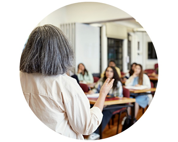 A teacher with gray hair speaks to a classroom of attentive students seated at desks in a learning environment.